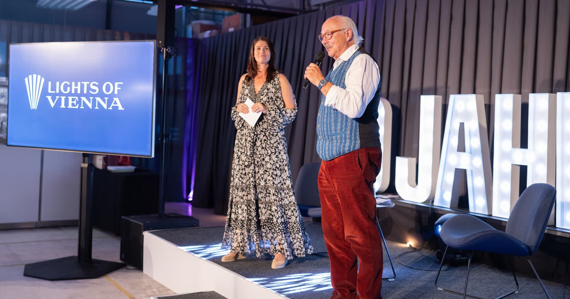 Speaker on stage during the Lights of Vienna anniversary celebration with illuminated logo in the background