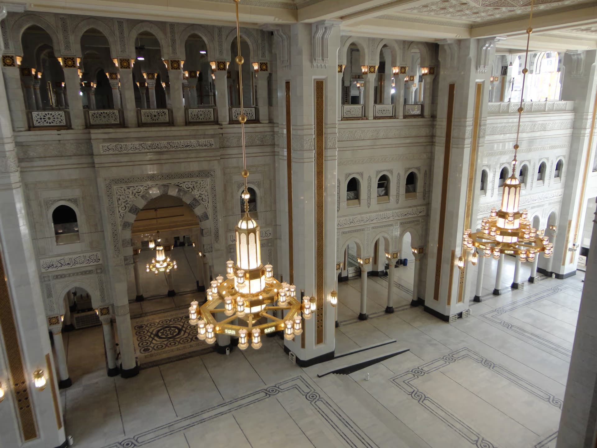Interior view of the Holy Mosque in Makkah featuring marble arches, Arabic calligraphy and grand golden chandeliers